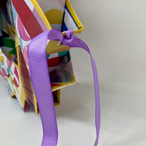 Pu&ograve; includere: Una scatola regalo colorata con motivo geometrico e un nastro viola legato intorno alla parte superiore.