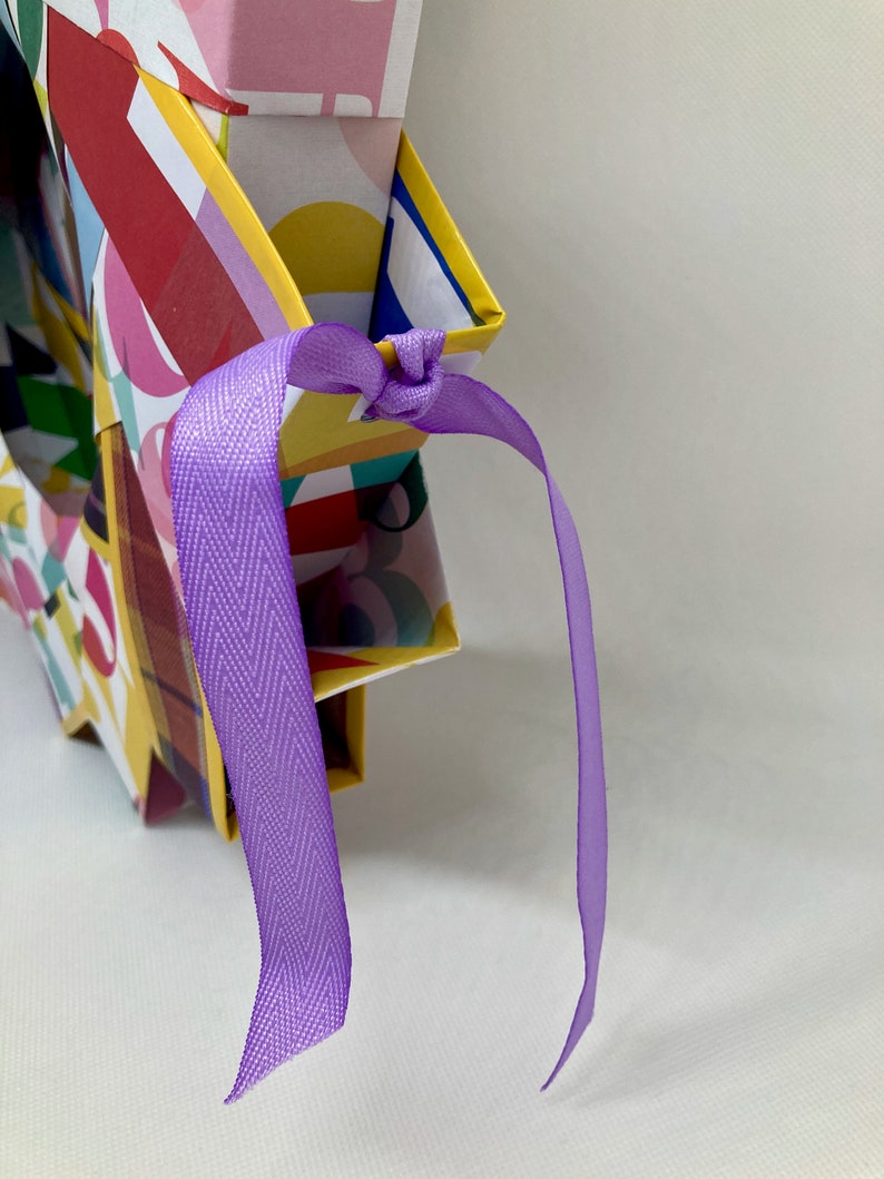 Pu&ograve; includere: Una scatola regalo colorata con motivo geometrico e un nastro viola legato intorno alla parte superiore.