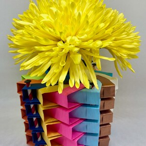 Pu&ograve; includere: Un vaso di origami colorato fatto di carta piegata in tonalit&agrave; di rosa, blu, giallo e arancione. Il vaso &egrave; pieno di un crisantemo giallo brillante.