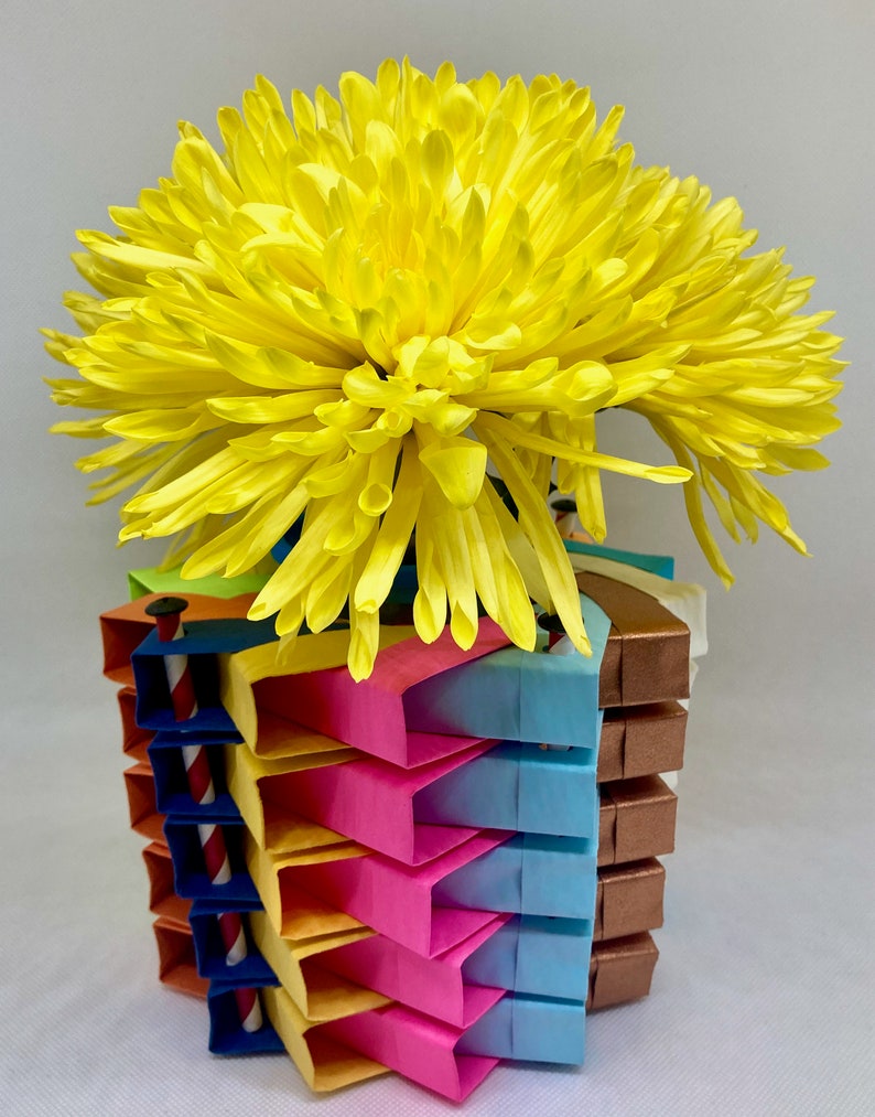 Pu&ograve; includere: Un vaso di origami colorato fatto di carta piegata in tonalit&agrave; di rosa, blu, giallo e arancione. Il vaso &egrave; pieno di un crisantemo giallo brillante.