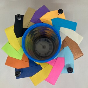 Pu&ograve; includere: Un bicchiere di carta blu con un motivo geometrico colorato &egrave; circondato da un anello di strisce di carta colorate, ciascuna con una vite fissata. Le strisce di carta sono disposte in un motivo circolare, creando un design decorativo.