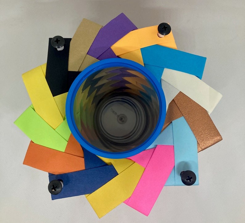 Pu&ograve; includere: Un bicchiere di carta blu con un motivo geometrico colorato &egrave; circondato da un anello di strisce di carta colorate, ciascuna con una vite fissata. Le strisce di carta sono disposte in un motivo circolare, creando un design decorativo.