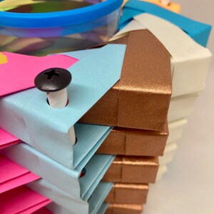 Pu&ograve; includere: Una pila di quadrati di carta origami colorati con un contenitore di plastica trasparente in cima. I quadrati sono disposti a spirale e sono tenuti insieme da viti nere. I colori dei quadrati sono rosa, blu, bianco e marrone.
