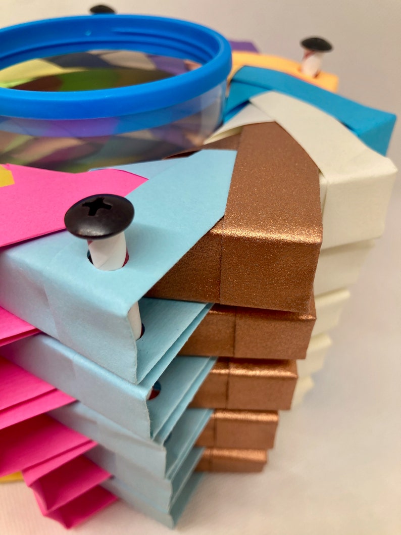 Pu&ograve; includere: Una pila di quadrati di carta origami colorati con un contenitore di plastica trasparente in cima. I quadrati sono disposti a spirale e sono tenuti insieme da viti nere. I colori dei quadrati sono rosa, blu, bianco e marrone.
