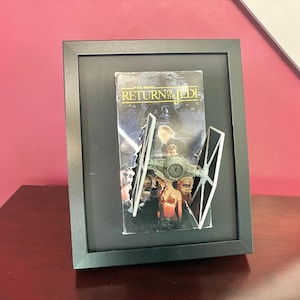 May include: Framed Star Wars: Return of the Jedi VHS tape with a 3D TIE fighter model. The black frame sits on a dark wood surface against a pink wall. The VHS cover features movie characters and the film title.