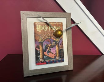 May include: Framed artwork featuring the book cover of "Harry Potter and the Sorcerer's Stone" by J.K. Rowling. A golden Snitch with silver wings is positioned above the book. The frame is a light gray color.