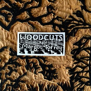 May include: A white sticker with black text that reads "WOODCUTS by Margot Torrey" on a brown wooden background with black lines.