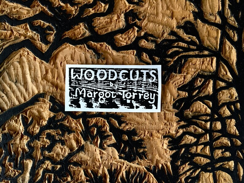May include: A white sticker with black text that reads "WOODCUTS by Margot Torrey" on a brown wooden background with black lines.