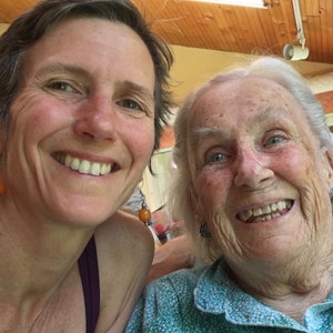 May include: Two women smiling at the camera. The woman on the left has short brown hair and is wearing a purple tank top. The woman on the right has white hair and is wearing a blue and green patterned shirt.