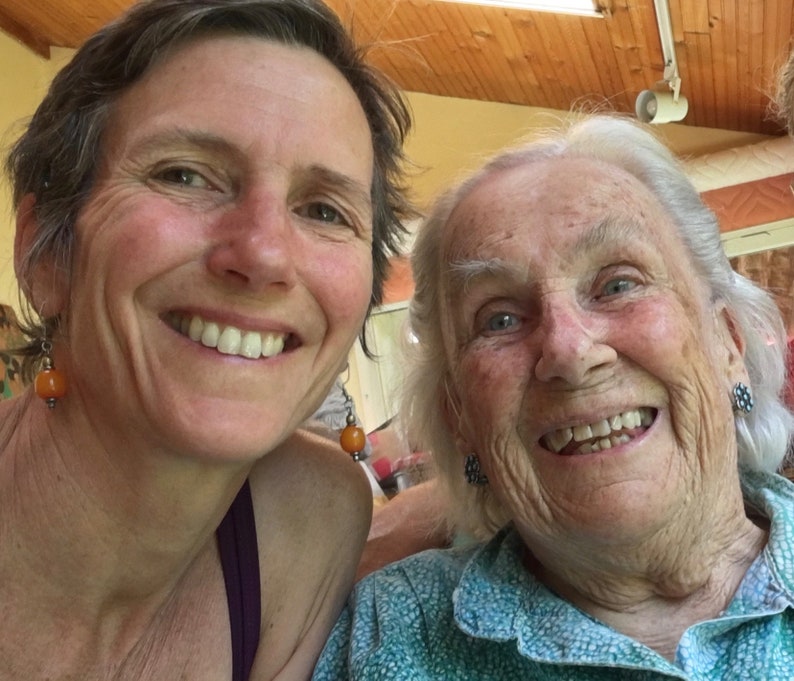 May include: Two women smiling at the camera. The woman on the left has short brown hair and is wearing a purple tank top. The woman on the right has white hair and is wearing a blue and green patterned shirt.