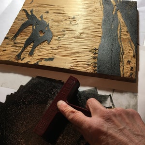 May include: A close-up of a wooden printing block with a black ink design of a creature. A hand is using a red rubber brayer to spread ink on a black surface.