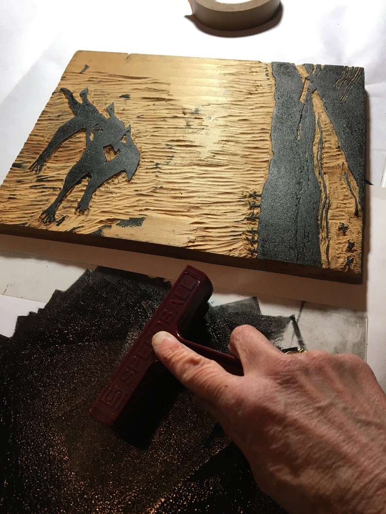 May include: A close-up of a wooden printing block with a black ink design of a creature. A hand is using a red rubber brayer to spread ink on a black surface.