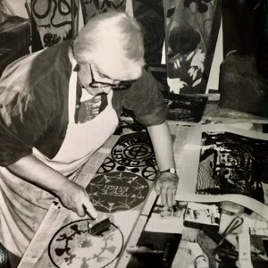 May include: A person in a white apron is working on a black and white print with a brush. The print is circular and has the words "Dance!" written on it. The person is surrounded by other prints and tools.