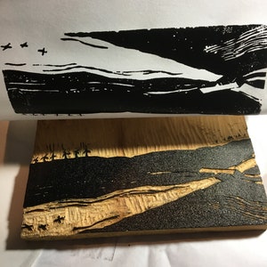 May include: A black and white woodblock print of a landscape with a river and mountains. The print is on a piece of wood.