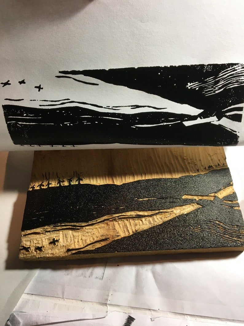 May include: A black and white woodblock print of a landscape with a river and mountains. The print is on a piece of wood.