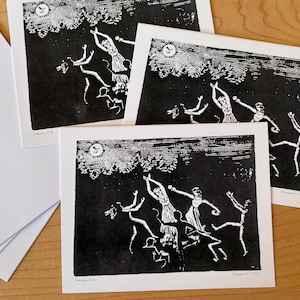 May include: Black and white linocut print of a group of figures dancing under a full moon. The print is on white paper and has a handwritten signature in the bottom right corner.