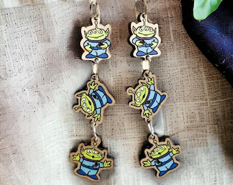 May include: A pair of dangle earrings featuring three wooden charms of the alien character from the Toy Story franchise. Each charm is painted with a green face, three eyes, and a blue body. The charms are connected by silver jump rings.