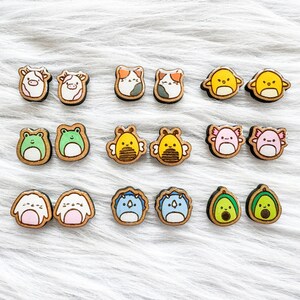 May include: A set of 10 pairs of wooden earrings featuring various cute animal and food designs. The earrings are painted in bright colors, including yellow, blue, green, pink, and brown. The designs include a cow, a cat, a chick, a frog, a bee, an axolotl, a penguin, and an avocado.