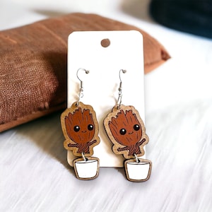 May include: A pair of wooden earrings featuring a cartoon image of a brown character with a pot on its head. The character is smiling and has large eyes. The earrings are hanging from silver hooks.