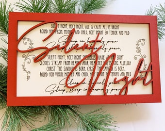 May include: A red wooden frame with a white background and the lyrics to the Christmas carol "Silent Night" printed in black. The words "Silent Night" are cut out of the frame and are red.