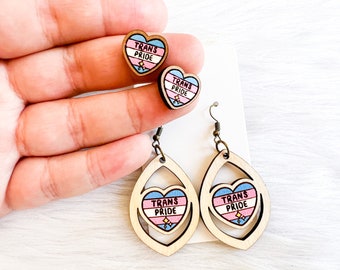 May include: A pair of wooden earrings with a trans pride heart design. The earrings are shaped like teardrops and have a smaller heart cutout in the center. The hearts are painted with the colors of the transgender pride flag and the words "Trans Pride" are written on them.