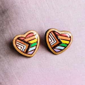 May include: A pair of wooden heart-shaped earrings with a rainbow geometric design. The earrings are painted with shades of pink, orange, yellow, green, blue, and purple.