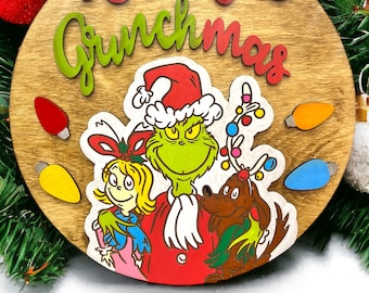 May include: A round wooden sign with the words "Merry Grinchmas" in red and green lettering. The sign features a green Grinch character wearing a red Santa hat, a blonde girl, and a brown dog.