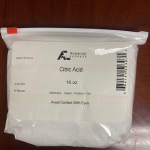 以下が含まれることがあります： クエン酸が入った透明なビニール袋。ラベルには「Citric Acid」と「16 oz」と書かれています。袋には「Avoid Contact With Eyes」と「Vegan, Paraben-Free」の文字も含まれています。