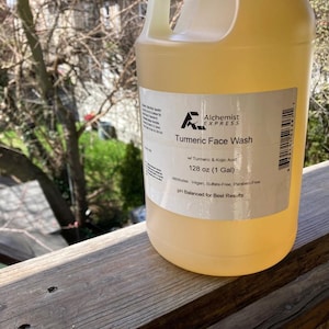 May include: A gallon jug of Turmeric Face Wash by Alchemist Express. The label is white with black text and a barcode. The text reads "Turmeric Face Wash with Turmeric & Kojic Acid 128 oz (1 Gal) Attributes: Vegan, Sulfate Free, Paraben Free pH Balanced for Best Results".
