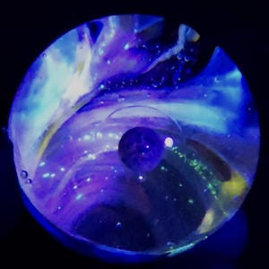May include: A glass marble with a galaxy design. The marble features swirling blues, purples, and white, with a small, dark blue sphere at the center. The background is black.