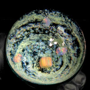 May include: A round, glass art piece with an internal design resembling a galaxy. The piece features swirling patterns in shades of blue, green, and white, with several iridescent, rectangular and spherical inclusions. The background is black.