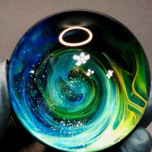 May include: A spherical glass paperweight featuring a swirling nebula pattern in shades of deep blue, vibrant green, and yellow. The interior contains small white flower-shaped inclusions and tiny sparkling glitter particles suspended within the clear glass.