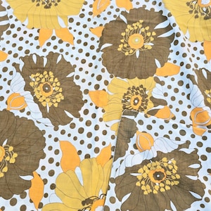 May include: A fabric featuring a repeating floral design. Large brown and yellow flowers with orange accents are set against a white background with brown polka dots. The fabric has a vintage aesthetic.