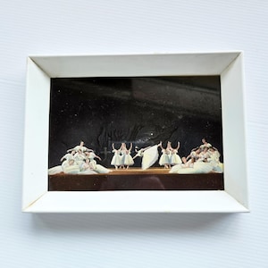 Puede incluir: Una obra de arte enmarcada que representa una escena de ballet. La obra muestra bailarinas con vestidos y tutús blancos en un escenario con un fondo oscuro. El marco es blanco.