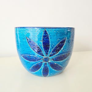 May include: A turquoise ceramic bowl with a dark blue floral design. The bowl has a textured surface and a rounded shape. The flower design is centered on the bowl and has six petals. The bowl is likely used for decorative purposes.