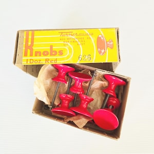 Puede incluir: Una caja de pomos rojos para armarios. La caja amarilla tiene la palabra "Knobs" en letras grandes y el texto "1 Doz. Red". La caja está abierta, revelando seis pomos rojos con tornillos.