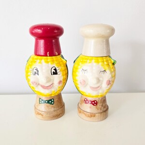 Może przedstawiać: Para ceramicznych solniczek i pieprzniczek w stylu vintage. Jedna solniczka ma czerwoną czapkę, druga białą czapkę kucharską. Obie mają żółte twarze z uśmiechniętymi wyrazami twarzy, różowymi policzkami i muszkami. Podstawy są jasnobrązowe.