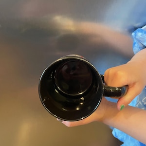 May include: A black ceramic coffee mug with a handle. The mug is held up, showing the inside of the mug. The mug is empty and has a glossy finish.