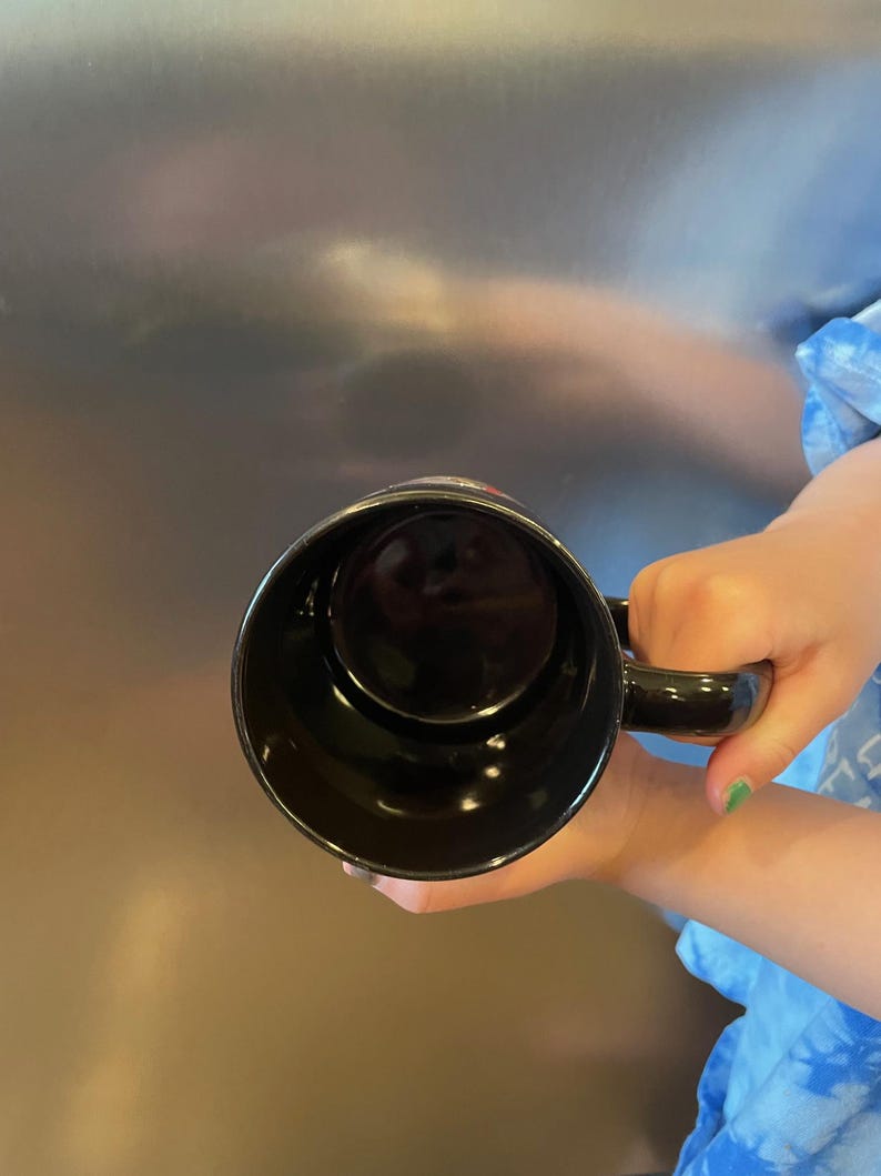 May include: A black ceramic coffee mug with a handle. The mug is held up, showing the inside of the mug. The mug is empty and has a glossy finish.