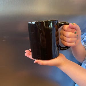 May include: A glossy black ceramic mug with a curved handle. The mug is held in a person's hand, showcasing its smooth, dark surface. The mug's design is simple and classic, suitable for coffee, tea, or other beverages.