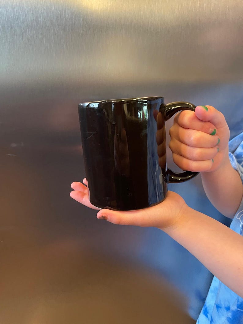 May include: A glossy black ceramic mug with a curved handle. The mug is held in a person's hand, showcasing its smooth, dark surface. The mug's design is simple and classic, suitable for coffee, tea, or other beverages.