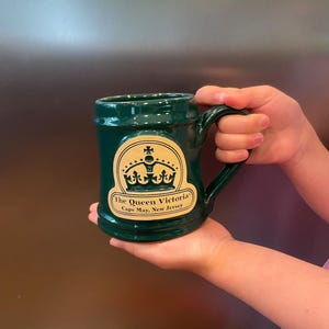 May include: A dark green ceramic mug featuring a tan, arched emblem on the front. The emblem displays a black crown graphic above the text "The Queen Victoria" and "Cape May, New Jersey". The mug has a sturdy handle and a smooth, glossy finish.