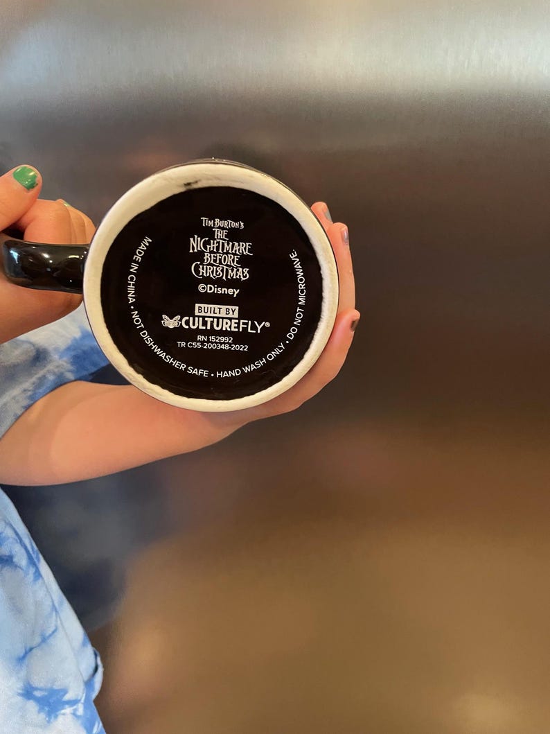 May include: Black ceramic mug featuring the text "Tim Burton's The Nightmare Before Christmas" and the Disney logo. The mug is built by Culturefly and is marked as hand wash only and not microwave safe.