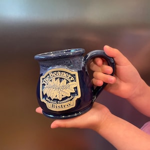 May include: A dark blue ceramic mug with a lighter blue rim and a handle. The front features a raised, cream-colored emblem with the text "The Beanery" above a coffee plant illustration and "Windsor, Connecticut Bistro" printed below.
