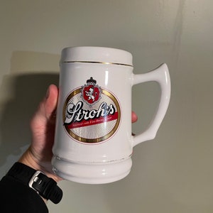 May include: White ceramic beer stein with a gold rim and handle. The stein features the Stroh's logo in a circular design with red, gold, and white colors. The text "America's only Fire-Brew Beer" is also visible.