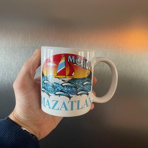 May include: White ceramic mug featuring a colorful design of a sailboat, dolphins, and the words "Mexico" and "Mazatlan." The design includes a sunset with red, yellow, and blue hues. The mug has a curved handle.