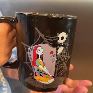 May include: Black ceramic mug featuring a colorful illustration of Sally and Jack Skellington from The Nightmare Before Christmas. Jack is holding a red rose, and Sally is seated. The mug has a black handle.
