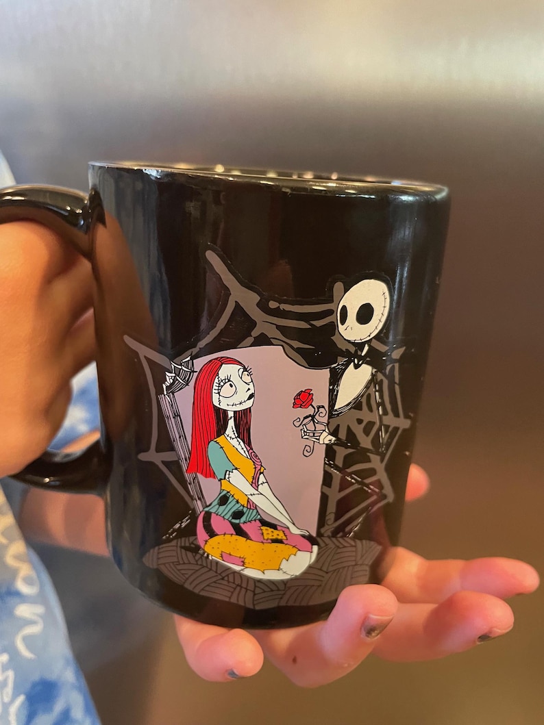 May include: Black ceramic mug featuring a colorful illustration of Sally and Jack Skellington from The Nightmare Before Christmas. Jack is holding a red rose, and Sally is seated. The mug has a black handle.