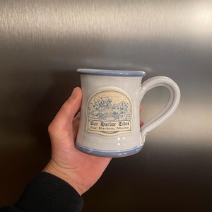 May include: A light blue ceramic mug with a dark blue rim and base. The front features a cream-colored emblem with a blue line drawing of a house and the text Bar Harbor Tides, Bar Harbor, Maine. The mug has a curved handle on the right side.