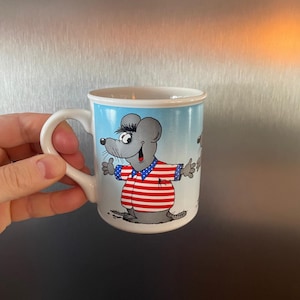 Puede incluir: Taza de cerámica blanca con un diseño de ratón de dibujos animados. El ratón lleva una camiseta a rayas rojas y blancas y una chaqueta azul con estrellas blancas. La taza tiene un fondo de cielo azul y un asa blanca.